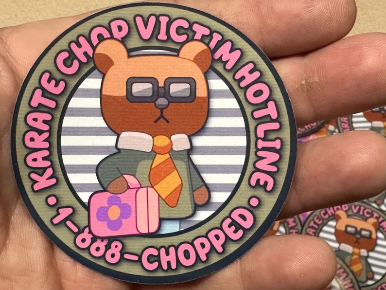 Puede incluir: Una pegatina redonda con un oso de dibujos animados que lleva gafas y corbata. La pegatina tiene el texto "KARATE CHOP VICTIM HOTLINE &bull; 1-888-CHOPPED" en letras rosas alrededor del borde.
