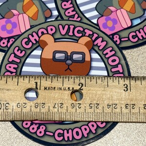 Puede incluir: Un oso marr&oacute;n con gafas sobre un fondo a rayas rosa y blanco. El oso sostiene una maleta rosa con una flor encima. El texto alrededor del oso dice "Rate Chop Victim Hot 888-Chopped".