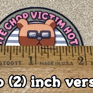 Puede incluir: Una pegatina redonda de 5 cm de di&aacute;metro con un oso de dibujos animados que lleva gafas. El oso est&aacute; rodeado de un c&iacute;rculo a rayas gris y blanco con el texto "ATE CHOP VICTIM HOT" en letras rosas.