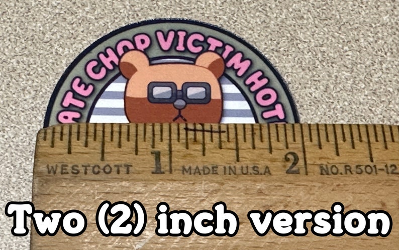 Puede incluir: Una pegatina redonda de 5 cm de di&aacute;metro con un oso de dibujos animados que lleva gafas. El oso est&aacute; rodeado de un c&iacute;rculo a rayas gris y blanco con el texto "ATE CHOP VICTIM HOT" en letras rosas.