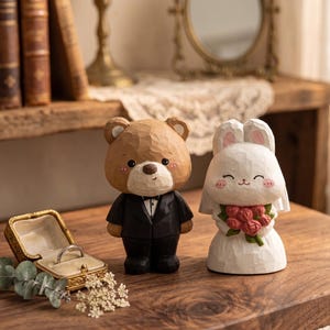 May include: A wooden bear groom and a rabbit bride figurine, with a ring box. The bear wears a black tuxedo, and the rabbit holds a bouquet of red roses. The ring box is gold with a ring inside.