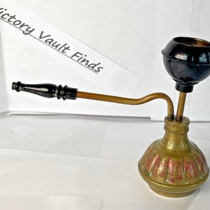 May include: An antique brass water pipe with a black bowl and mouthpiece. The pipe has a bulbous base with red and gold etched designs. The text "Victory Vault Finds" is in the background.
