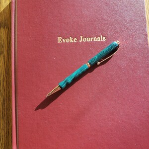 May include: A burgundy journal with the gold text "Evoke Journals" and a green and gold pen. The pen is lying on top of the journal. The journal has a red ribbon bookmark.