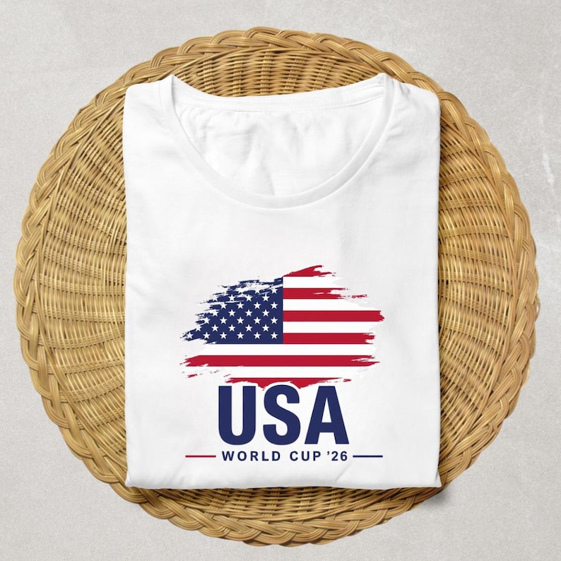 May include: White t-shirt featuring a stylized American flag design in red, white, and blue. The word "USA" is printed in navy blue below the flag, with "WORLD CUP '26" underneath in a smaller font. The shirt is folded on a woven wicker surface.