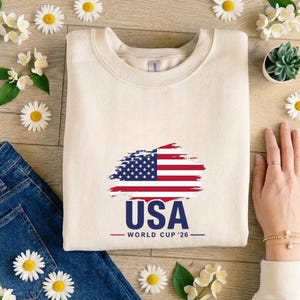 May include: Cream-colored sweatshirt with a distressed American flag design and the text "USA WORLD CUP '26" in navy blue and red. The sweatshirt is paired with blue jeans and floral accents.