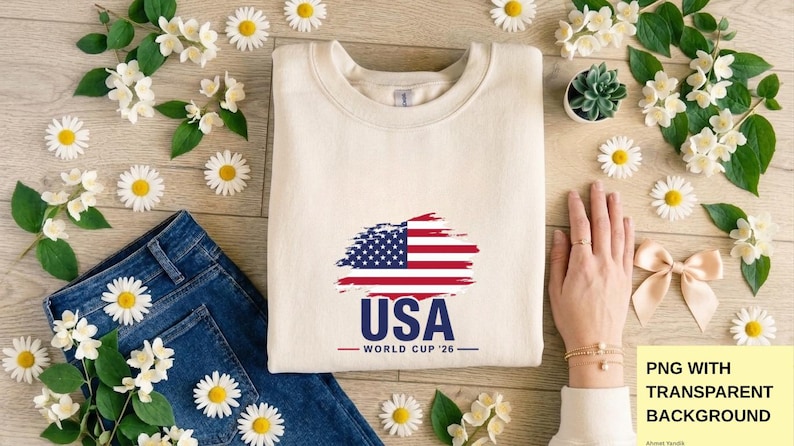 May include: Cream-colored sweatshirt with a distressed American flag design and the text "USA WORLD CUP '26" in navy blue and red. The sweatshirt is paired with blue jeans and floral accents.