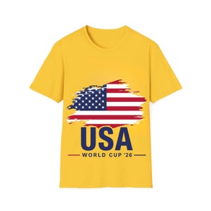 May include: A yellow t-shirt featuring a graphic of the American flag with a brushstroke effect. Below the flag, the word "USA" is printed in navy blue, with "WORLD CUP '26" underneath in a smaller font.