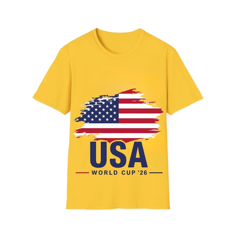 May include: A yellow t-shirt featuring a graphic of the American flag with a brushstroke effect. Below the flag, the word "USA" is printed in navy blue, with "WORLD CUP '26" underneath in a smaller font.