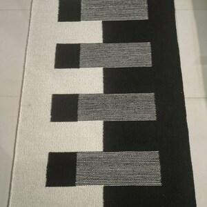 Puede incluir: Una alfombra rectangular con un diseño dividido, una mitad en blanco sólido y la otra en negro sólido. Cuatro bloques rectangulares horizontales se extienden desde el lado blanco hacia el lado negro, alternando con franjas texturizadas.