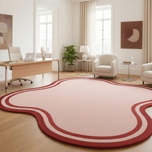 May include: An irregular, cloud-shaped area rug featuring a soft pink center surrounded by two distinct, concentric dark red border lines. The rug is positioned on a light-colored wooden floor in a room with white walls and neutral furniture.