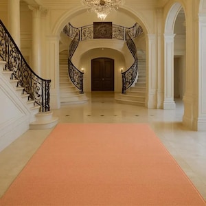 May include: A rectangular area rug in a solid, muted peach color with a clean, finished edge. The rug features a flat, uniform texture and is positioned centrally on a light-colored, polished stone floor within a grand, open interior space.