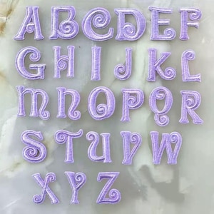 May include: A complete set of twenty-six individual embroidered alphabet letters from A to Z. Each letter is rendered in a light lavender thread with a distinct swirling decorative detail integrated into the font design of every character.