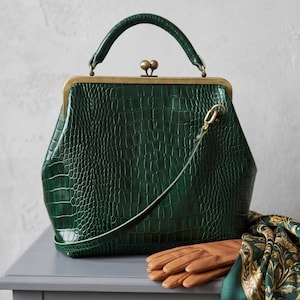 May include: A dark green handbag with a crocodile-embossed texture, featuring a brass-toned metal frame, a top handle, and a kiss-lock closure. The bag includes a thin shoulder strap and is positioned next to tan leather gloves and a patterned scarf.