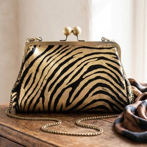 May include: A structured clutch bag featuring a black and metallic gold zebra stripe pattern. The bag has a brass-toned metal frame with a dual ball clasp closure and a matching thin metal chain shoulder strap attached to the sides.