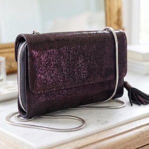 Pode incluir: Uma clutch roxa escura com uma corrente prateada e uma borla. A clutch tem um acabamento texturizado e brilhante e um fecho de correr. Está pousada numa superfície de mármore branco.