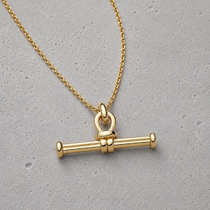 Pode incluir: Um colar dourado com fecho de barra e anel. O colar apresenta uma corrente delicada e um pendente de barra simétrico e polido com extremidades arredondadas. O fecho é um design distinto em forma de T, criando um visual moderno e elegante.