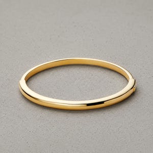 May include: A gold-colored bangle bracelet. The bracelet is a simple, rounded design with a smooth, polished finish. The jewelry is set against a gray background.