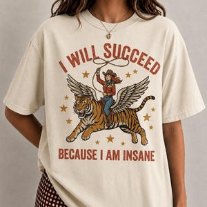 May include: A cream-coloured t-shirt featuring a graphic print of a person in a cowboy hat riding a winged tiger while holding a lasso. The text "I WILL SUCCEED" is arched above the illustration, and "BECAUSE I AM INSANE" is printed below in red.