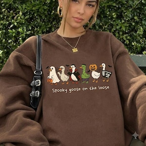 Puede incluir: Una sudadera marrón de cuello redondo con un estampado gráfico de seis gansos vestidos con varios disfraces de Halloween, incluyendo un fantasma, momia, vampiro, monstruo, calabaza y esqueleto, con el texto Spooky goose on the loose debajo.