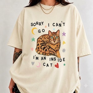 May include: A cream-colored short-sleeve t-shirt featuring a graphic of a brown tabby cat. The text reads "SORRY I CAN'T GO I'M AN INSIDE CAT" in black letters, surrounded by colorful small stars, hearts, and crescent moon shapes.