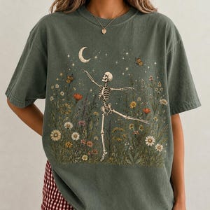 May include: A dark olive green short-sleeved t-shirt featuring a graphic print of a skeleton dancing in a field of wildflowers. The design includes a crescent moon, scattered stars, and butterflies, all rendered in muted, earthy tones across the chest.