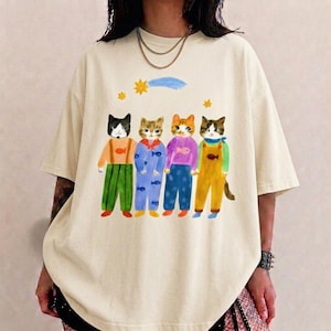 Può includere: Una t-shirt a maniche corte color crema con un'illustrazione grafica di quattro gatti in piedi che indossano abiti colorati. I gatti sono raffigurati con vari outfit, tra cui salopette e maglioni con motivi a pesci, sotto una stella gialla e una cometa blu.