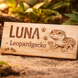 Könnte beinhalten: Holztafel mit dem eingravierten Namen "LUNA" und "- Leopardgecko". Rechts ist ein Cartoon-Leopardgecko abgebildet. Pfotenabdrücke und Blätter sind ebenfalls auf der Tafel eingraviert. Die Tafel liegt auf sandigem Untergrund.