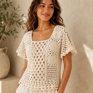 May include: A cream-colored crochet top featuring a square neckline, short flared sleeves, and a complex geometric pattern of interconnected circular and square motifs. The garment is crafted with an open-knit design and a scalloped hemline.