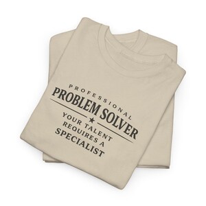 May include: A beige short-sleeved t-shirt featuring black text that reads PROFESSIONAL PROBLEM SOLVER, with a small star icon, followed by the text YOUR TALENT REQUIRES A SPECIALIST. The shirt is folded and layered on top of another identical beige t-shirt.