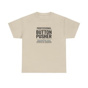 Op de afbeelding: Een zandkleurig t-shirt met korte mouwen en zwarte tekst op de borst met de tekst PROFESSIONAL BUTTON PUSHER, met daaronder kleinere tekst met TESTED BY PLC en APPROVED BY ENGINEERS, gescheiden door horizontale lijnen en een tandwielicoon.