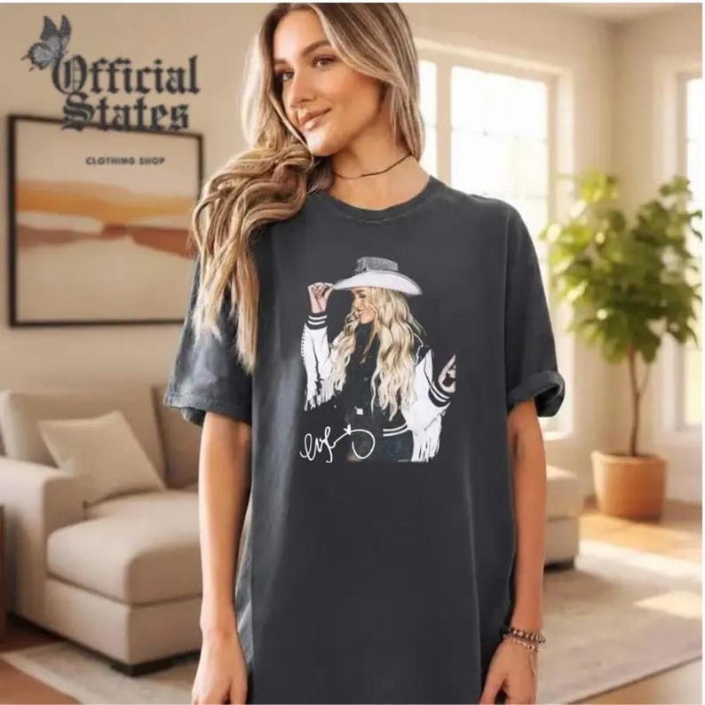 Puede incluir: Camiseta gris oscuro con un gr&aacute;fico de una mujer con sombrero y chaqueta, con una firma debajo. Las mangas est&aacute;n enrolladas. El texto "Official States Clothing Shop" es visible en el fondo.