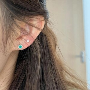 May include: Two stud earrings worn in the earlobe. The larger earring features a central round green gemstone surrounded by a halo of small clear stones. The smaller earring is a single, tiny clear stone set in a simple metal prong setting.