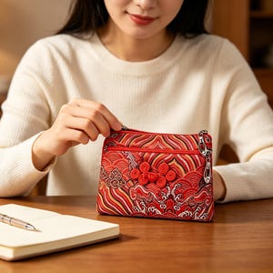 May include: Red and gold embroidered clutch with a zipper closure. The clutch features an intricate floral and wave pattern. The bag is held by a person wearing a white sweater. A notebook and pen are on the table.
