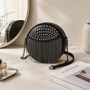 May include: Black round crossbody bag with silver studs and fringe. The bag has a chain strap and a black leather strap. A mirror and a stack of books are in the background.