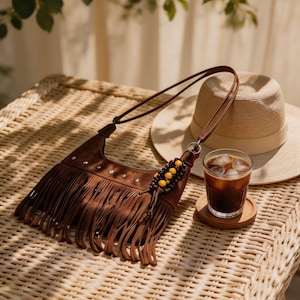 May include: A brown suede fringe bag with a long strap, embellished with small silver studs and a beaded accent. A straw hat and a glass of iced coffee sit on a wicker table, creating a relaxed, bohemian vibe.