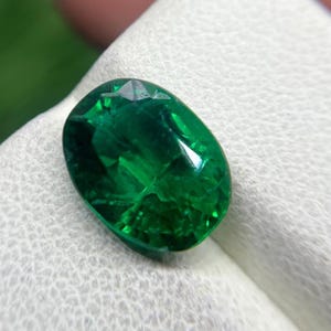 May include: An oval-cut gemstone with a deep, vibrant green colour and a faceted surface that reflects light. The stone displays internal clarity and natural inclusions, showcasing a brilliant, saturated emerald green hue throughout its structure.