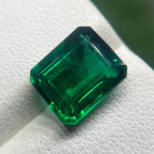 May include: A loose emerald-cut gemstone with a deep, vibrant green color and a faceted surface that reflects light. The stone has a rectangular shape with clipped corners, showcasing internal clarity and a polished, glassy texture.