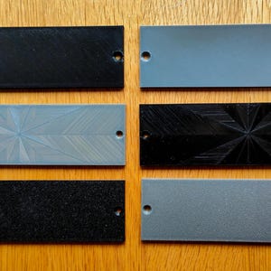 May include: A collection of rectangular, textured panels in various colors, including black, gray, and blue. Some panels feature patterned designs, and all have a single hole. The panels are arranged on a wooden surface.