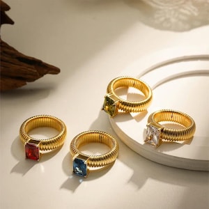 Könnte beinhalten: Vier goldfarbene Ringe mit quadratischen Edelsteinen in Rot, Blau, Grün und Klar. Die Ringe haben ein geripptes Banddesign. Die Edelsteine sind in einer goldenen Lünette gefasst. Die Ringe werden auf einer weißen Oberfläche präsentiert.