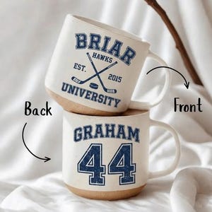 May include: Two white ceramic mugs with a tan base are stacked. The top mug displays "BRIAR HAWKS UNIVERSITY" with crossed hockey sticks and the text "EST. 2015" in blue. The bottom mug has "GRAHAM 44" in blue.