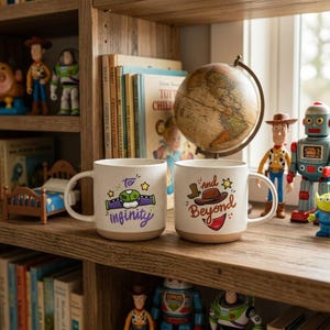 May include: Two off-white ceramic mugs with speckled textures. The left mug features a green and purple space ranger chest plate graphic with the text To Infinity. The right mug features a brown cowboy hat and red bandana graphic with the text and Beyond.
