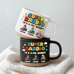 May include: Two ceramic mugs stacked on top of each other. The top mug is white with a tan base and a black handle. The bottom mug is black with a tan base and a black handle. Both mugs have the text "SUPER DADDIO" and "IT'S A-ME, DADDY!" with cartoon characters.