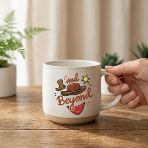 May include: A speckled white ceramic mug featuring a brown cowboy hat, a small boot, a yellow star, and a red bandana graphic. The text "And Beyond" is printed in a brown script font on the front of the mug, which has a sturdy handle.