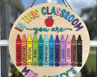 Könnte beinhalten: Runder Holzklassenschild mit der Aufschrift "In This Classroom You Are" und einer Liste positiver Eigenschaften. Das Schild zeigt einen roten Apfel und bunte Buntstifte, mit dem Namen "Mrs. Ashley's" am unteren Rand.
