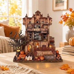 May include: A decorative model of The Hollywood Tower Hotel featuring a weathered facade, illuminated windows, and a spooky tree with a jack-o'-lantern face. The base includes small ghost figures, pumpkins, and a sign reading The Twilight Zone Tower of Terror.