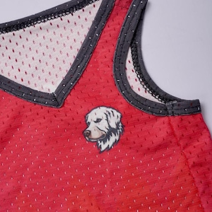 May include: A red sleeveless mesh tank top featuring a V-neckline with dark gray trim. The garment is constructed from a perforated athletic fabric and displays a small, detailed graphic illustration of a white dog head on the front chest area.