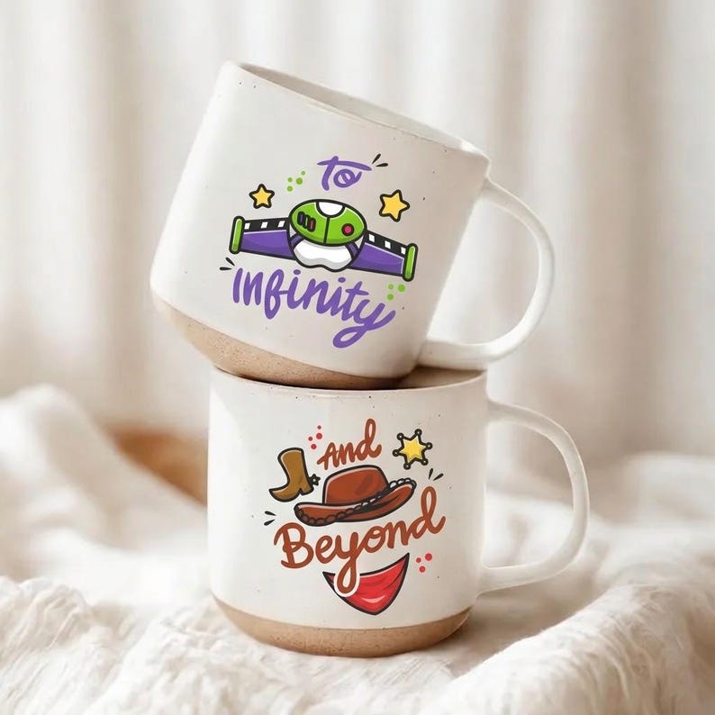 May include: Two stacked ceramic mugs with a speckled off-white finish. The top mug features a space ranger chest plate graphic with the text "to infinity". The bottom mug displays a cowboy hat, boot, and bandana graphic with the text "And Beyond".
