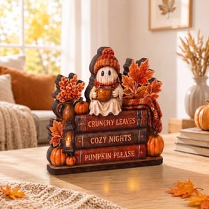 Puede incluir: Una figura decorativa de mesa que muestra un fantasma con gorro y bufanda de punto naranja, sosteniendo una taza. El fantasma se sienta sobre tres libros apilados con las etiquetas CRUNCHY LEAVES, COZY NIGHTS y PUMPKIN PLEASE, junto a calabazas.