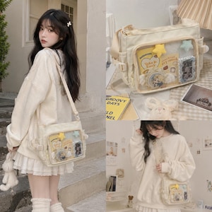 Puede incluir: Bolso mensajero color crema con un bolsillo frontal transparente que muestra objetos decorativos. El bolso tiene una correa larga y se lleva al hombro. Contenido: un libro amarillo, decoraciones en forma de estrella y otros objetos pequeños.