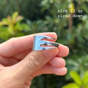 May include: A silver-colored ring crafted from a fork, featuring three tines. The ring is held in a hand against a blurred green background. Text on the image reads "size 12 or sized down."